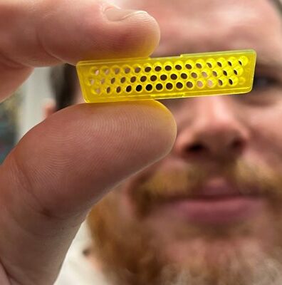 Ben Dopp holding a small yellow micro 3D printed connector part with three rows of tiny through-holes